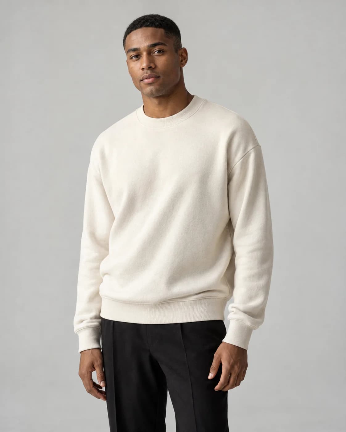 Adult male model wearing an ivory sweatshirt and black trousers in a gray studio